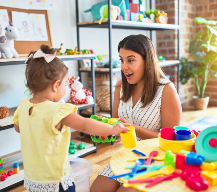 Young Beautiful Teacher And Toddler Playing With Dishes, Cutlery And Cups Toy On The Table At Kindergarten