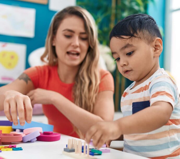 Teacher And Toddler Playing With Geometry Blocks Sitting On Table At Kindergarten