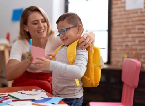 Teacher And Toddler Cutting Paper Wearing Backpack At Kindergarten