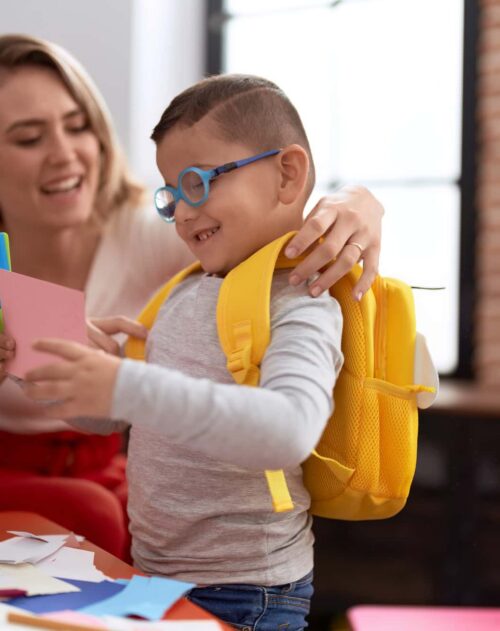 Teacher And Toddler Cutting Paper Wearing Backpack At Kindergarten