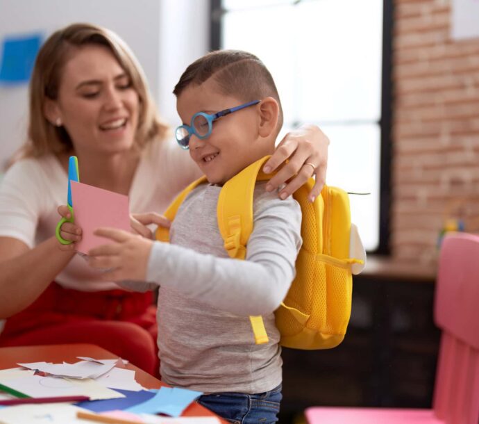 Teacher And Toddler Cutting Paper Wearing Backpack At Kindergarten